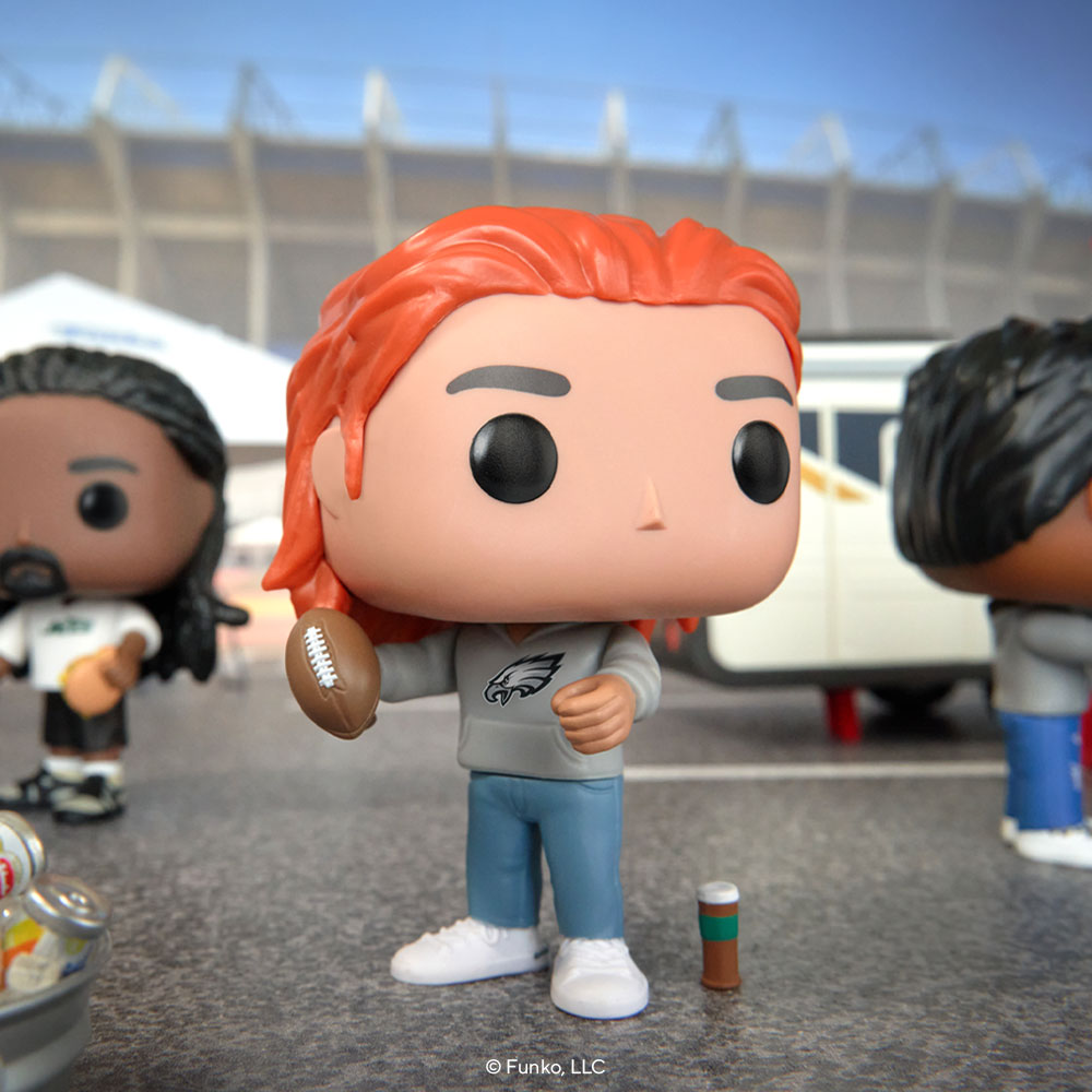 This lifestyle photo shows a red-headed POP! Yourself Eagles fan in a hoodie with the NFL logo, holding a football. They're just outside a stadium with other POP! Yourself football fans. Toy cars are in the background to imitate a tailgate party of sorts.